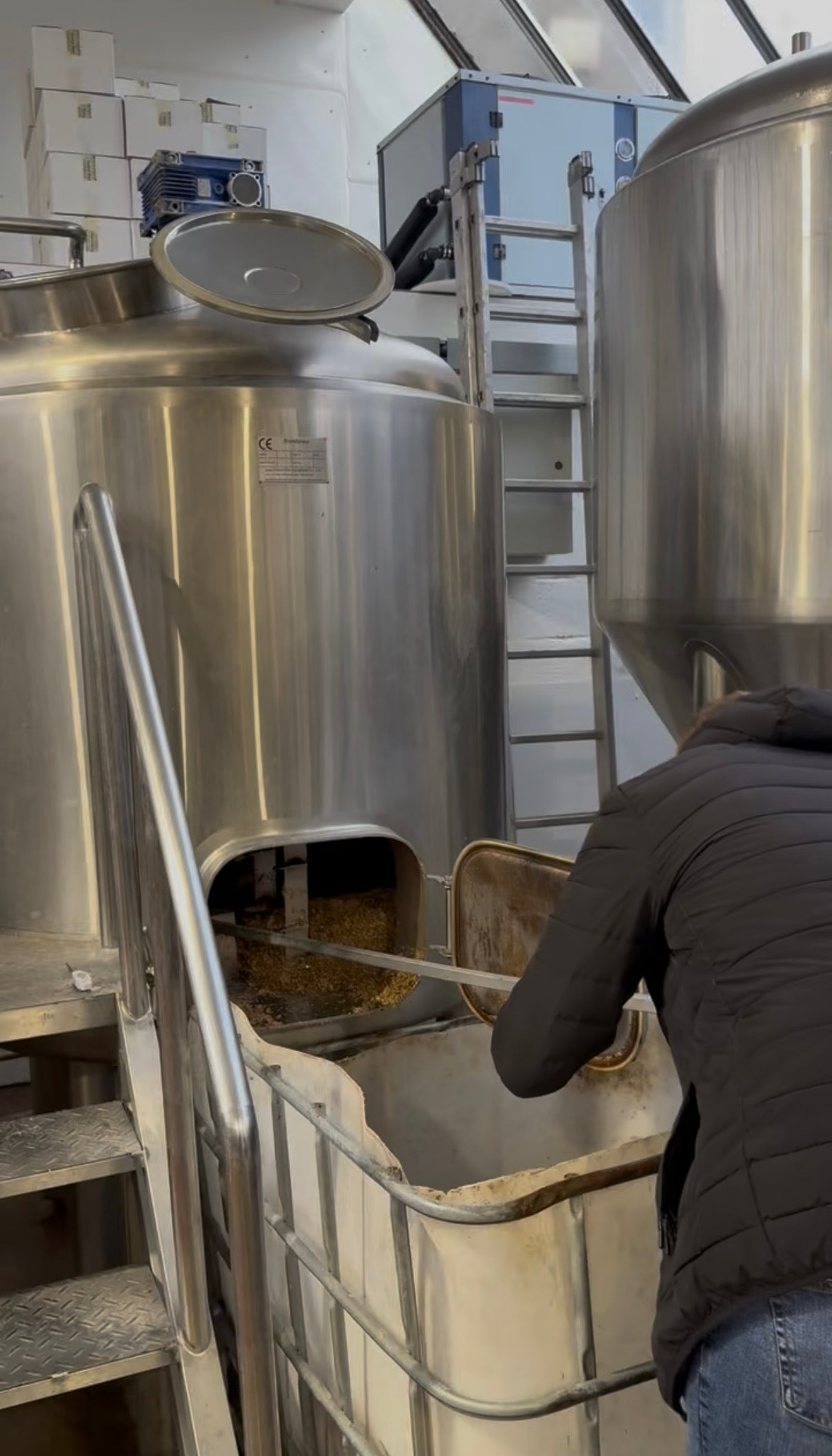 Person working in a brewery with large metal tanks and equipment.